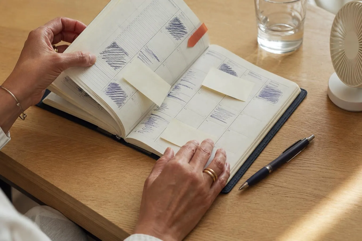 Gros plan sur un agenda papier ouvert couvert de notes irrégulières et de petits papiers repositionnables, sur une table claire, avec les mains d’une femme d’âge mûr